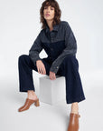 Woman sitting on a white cube wearing a dark denim outfit with a patterned shirt.