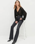 Woman wearing a black blouse and dark jeans sitting on a white stool against a white background