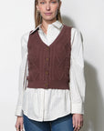 A woman modeling a brown cable-knit vest over a white and navy striped button-down shirt and blue jeans, standing against a plain light gray wall.