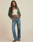 Woman wearing a green jacket, white top, and blue jeans on a beige background