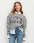 Woman wearing a gray sweatshirt with 'Favorite Daughter' text, light blue shirt, and jeans on a white background front view