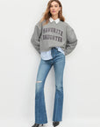 Woman wearing a gray 'Favorite Daughter' sweatshirt with a white shirt and blue jeans on a light gray background