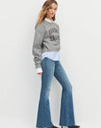 Woman wearing a gray sweatshirt with text, white shirt, and blue flared jeans on a white background side view