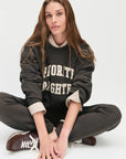 model wearing collegiate sweatshirt with brown pants and brown shoes sitting on the ground with a white background