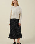 A woman stands against a plain beige studio background wearing a cream cable-knit sweater, a black midi skirt with small white polka dots, and black loafers.