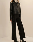 A model wearing a black cropped faux-leather jacket with structured shoulders, black wide-leg trousers, a white mock-neck top, and black pointed-toe ankle boots, standing against a warm off-white studio background.