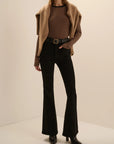 A model wearing a brown and black striped long-sleeve top, a tan open-front cardigan, black flared pants, and black ankle boots, standing against a plain beige studio background.