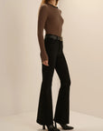 A model wearing a brown and black horizontally striped long-sleeve top, black flared pants, a black belt, and black pointed-toe heels, standing against a plain beige studio background.