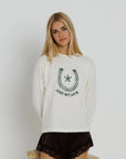 A blonde woman wearing a cream-colored long-sleeve sweatshirt with a dark green horseshoe and star graphic and the text 'JUST MYLUCK' on the front, paired with dark brown lace-trimmed shorts, against a plain light gray background.