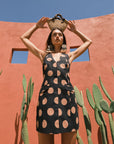 Woman in a black dress with tan polka dots holding a woven basket against a red wall with cacti.