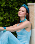 Woman in a blue dress and accessories sitting against a green tiled wall.