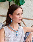 model with blue and white jewelry sitting with a couch and plant in the background
