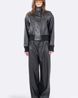 A model wearing a black faux-leather two-piece outfit consisting of a cropped high-collar jacket and matching wide-leg trousers, styled with gold-framed sunglasses against a light gray background.