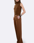 A woman modeling a brown sleeveless mock-neck knit top paired with matching wide-leg trousers, styled with sunglasses and bracelets against a light gray background.
