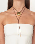 A close-up of a model wearing a brown double-strand cord necklace with a large, polished gold oval pendant and small gold bead accents at the ends, styled with a white strapless top.