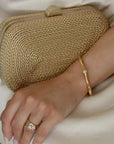 Gold clutch held by a hand with a gold ring and bracelet on a neutral background