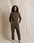 A woman modeling a matching two-piece tracksuit in a muted olive-brown color, consisting of a zip-up long-sleeve top and matching pants, standing against a draped off-white fabric backdrop.