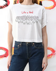 Person wearing a white t-shirt with 'Life is Hard' text and graphic design, standing against a colorful abstract background.