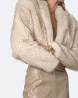 A model wearing a voluminous, shaggy faux fur jacket in a light cream/beige color, paired with a matching beige textured skirt, against a plain light gray background.