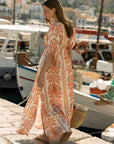 Woman in an orange and white patterned dress standing by a marina with boats in the background. back view