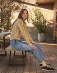A woman sits on a wooden bench on a sunlit deck, wearing a yellow and beige striped long-sleeve top, light blue jeans, white socks, and black sneakers.