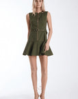 A blonde model wearing an olive green sleeveless mini dress with a front zipper, a matching belt with a gold buckle, and a flared skirt, paired with nude lace-up high heels against a white studio background.
