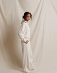 A woman wearing a matching white hooded sweatshirt and wide-leg sweatpants stands in profile against a draped cream fabric backdrop. side view
