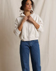 A woman wearing a cream-colored zip-up hoodie and blue jeans, standing against a neutral beige fabric backdrop.
