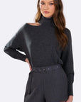 A woman modeling a dark gray knit turtleneck sweater with a shoulder cutout and matching dark gray pinstriped shorts with a belt, against a plain white studio background.