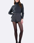 A model wearing a dark gray ribbed knit turtleneck sweater with a matching belt and pleated shorts, paired with sheer black tights and black pointed-toe heels, against a light gray studio background.
