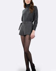 A woman modeling a dark gray ribbed knit turtleneck sweater and matching belted shorts, paired with sheer black tights and black pointed-toe heels, against a light gray studio background.