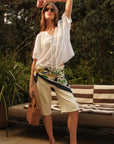 Woman in white outfit with floral pattern standing outdoors near a striped blanket.