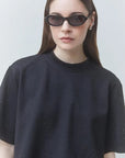 A woman wearing a black short-sleeve crewneck t-shirt with subtle silver rhinestone embellishments, styled with black cat-eye sunglasses against a light gray background.