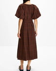 A woman viewed from the back wearing a brown and black plaid midi dress with puff sleeves and a tie waist, paired with black flat shoes, against a plain white studio background.