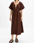 A woman modeling a brown and black plaid midi dress with puff sleeves, a deep V-neckline, and a matching fabric tie belt at the waist, paired with black strappy sandals against a plain white studio background.