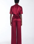 A model viewed from the back wearing a deep burgundy short sleeves top with a high collar, and wide leg pants, set against a plain white studio background.