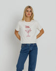 A woman wearing a white short-sleeve graphic t-shirt with a red 'TINI TIME' martini glass illustration on the front, paired with blue jeans, against a light gray studio background.