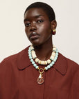 A model wearing Toro Earrings in Aubergine and a rust-brown oversized collared shirt and layered necklaces, including a prominent light blue and cream beaded necklace with a gold pendant, against a plain off-white background.