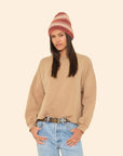 Woman wearing a beige sweater, striped beanie, and blue jeans on a beige background