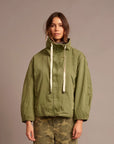 A woman wearing an olive green hooded jacket with cream drawstrings and olive camouflage pants, standing against a solid taupe background.