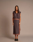 A woman stands against a plain taupe studio background wearing a sleeveless midi dress with a dark navy and rust red geometric pattern, ruffled cap sleeves, and a matching patterned belt.