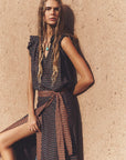 A woman models a sleeveless wrap-style midi dress with a dark navy and beige geometric pattern, featuring ruffled shoulders and a matching fabric tie belt at the waist, posed against a textured beige stucco wall.