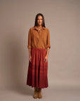 A woman stands against a plain warm beige studio background wearing a rust-brown button-up shirt and a long red skirt with a darker red gradient near the hem, paired with tan ankle boots.