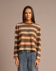 A woman modeling a cropped, long-sleeve knit sweater with horizontal stripes in brown, beige, and orange, paired with blue jeans, against a solid taupe background.