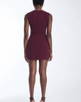 A woman viewed from the back wearing a sleeveless, knee-length burgundy dress with a structured silhouette and thin waist ties, paired with black strappy heels against a plain white studio background.