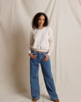 A woman stands against a draped cream fabric backdrop wearing a cream crewneck sweater, blue wide-leg jeans, and brown shoes.