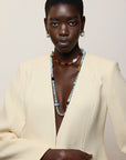 A model with short dark hair is wearing a cream-colored blazer, Mini Organic Hoops in Seafoam and layered beaded necklaces, including a prominent light blue beaded necklace, against a plain light beige background.