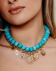 Close-up of a woman wearing a turquoise beaded necklace stacked with gold charm necklace