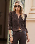 A woman wearing a dark brown sparkly knit cardigan with puffed shoulders, paired with dark pants and sunglasses, standing in front of a beige stone building.