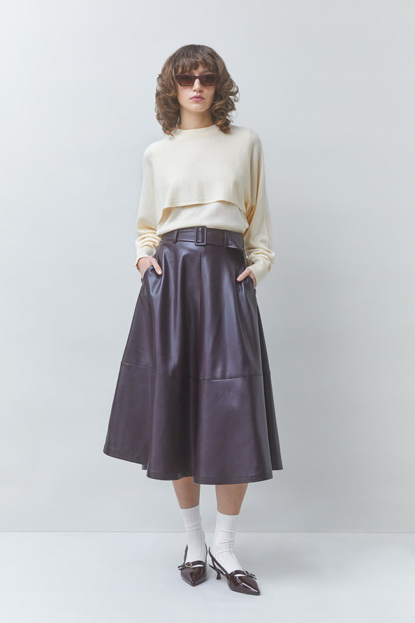 Person wearing a beige sweater, dark purple leather skirt, and white shoes on a plain background